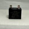 A black rectangular ADP250E106J-M2 10 uF 250 VAC capacitor (#410570-001) with two metal terminals on top, placed on a light gray surface. Technical details and certification logos are visible on the front.